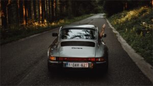 クラシックポルシェ911 カレラが森の道を走る。運転手が窓から手を出している後ろ姿