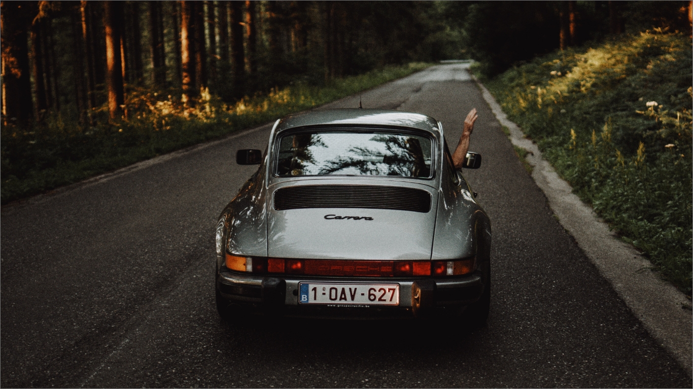 クラシックポルシェ911 カレラが森の道を走る。運転手が窓から手を出している後ろ姿