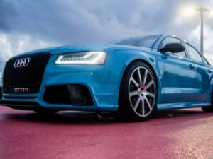 夕暮れ時の屋外で撮影された、青いAudi RSモデルの低角度ショット。LEDヘッドライトが点灯し、スポーティなホイールとフロントグリルが際立っている
